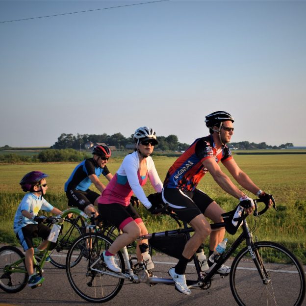 On this Friday this family is having fun and making memories, on a tandem bike with a tag-along (a.k.a. trailer cycle or trail-a-bike), riding across Iowa, on RAGBRAI 2018.